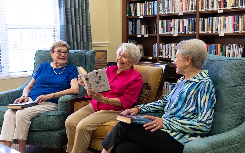 Three senior women reading books in the library at The Moorings of Arlington Heights