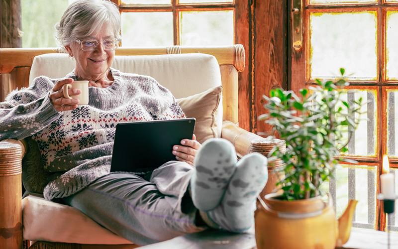 Senior woman sitting at home on armchair using digital tablet wearing a warm sweater and eyeglasses. Comfortable living room, wooden rustic windows