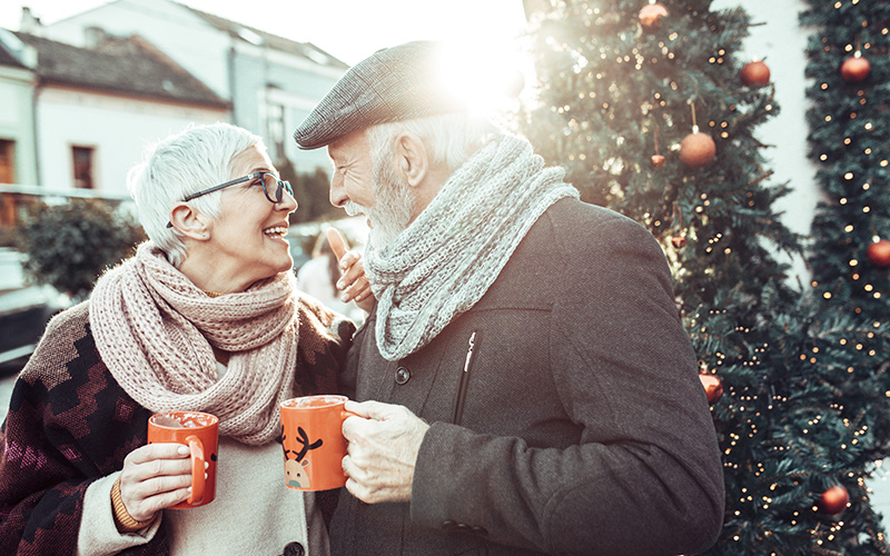 Senior couple with reindeer mugs filled with hot chocolate laughing in front of a decorated outdoor christmas tree.