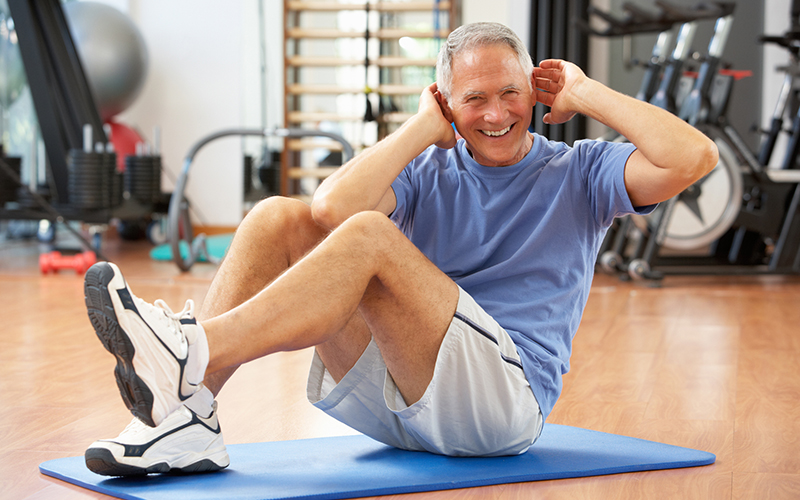 Senior Man Doing Sit Ups In Gym On Mat Smiling - clubs for seniors can help you stay active!