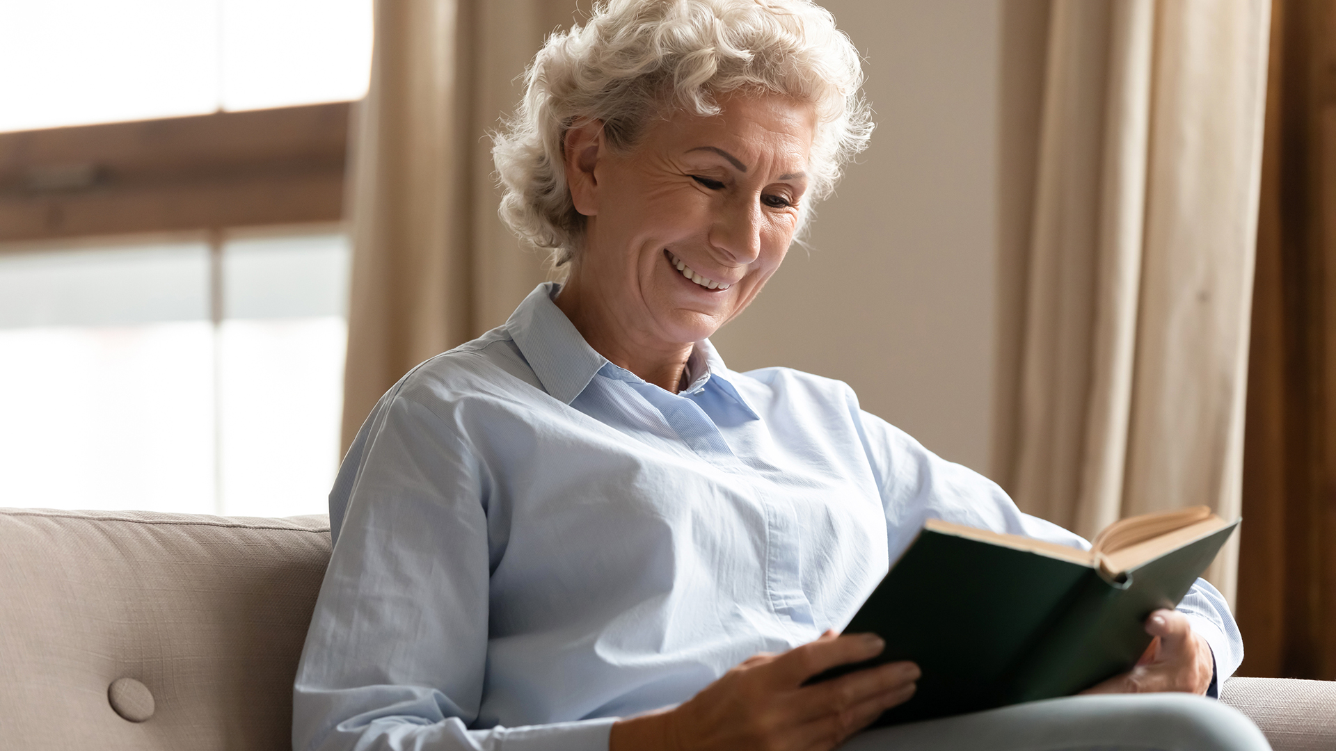 Smiling mature woman enjoy free time having good hobby pastime reading interesting literature holding book seated on comfy couch in living room at home alone